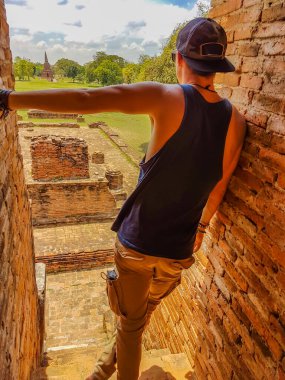 A a young man standing on one of the buildings of Ayutthaya Historical Park, Thailand, leaning on the side. UNESCO list. Ruins of an ancient city, built of bricks. beautiful and clear day.