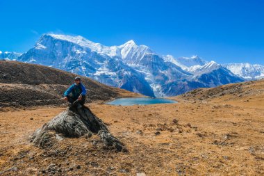 Man sitting on a rock, next to Ice lake, as part of the Annapurna Circuit Trek detour, Himalayas, Nepal. Annapurna chain in the back, covered with snow.  Dry grass, snowy peaks. Freedom and meditation