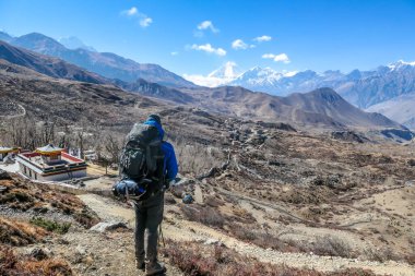 Young man carrying a huge backpack, wearing blue jacket looks down on the Muktinath city and the temple. Taken in Mustang Valley, Annapurna Circuit Trek, Himalayas, Nepal. Harsh and dry landscape.