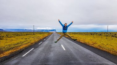 Young man wearing blue jacket jumps on the middle of an empty road. He spreads his limbs wide. Jump of joy, jump of happiness. Traveling to less traveled places. Sides of the road covered with grass.