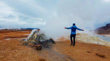 Young man wearing a blue walks into a steaming fumaroles emitting sulfuric gas with his arms wide spread to the sides. Power of planet Earth. Geothermal area displays large mud pools and steam vents