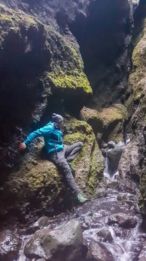 A young man wearing blue jacket tries to cross the spring without getting wet. He puts his leg down, supporting himself on the sides. The spring flows in a gorge. Steep and tall sides of the gorge.