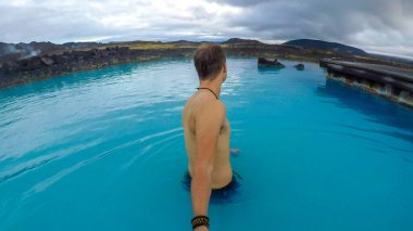 A young man holding a selfie stick and walking around the baby blue waters of Mvyatn Natural Bath. Nice and relaxing break while traveling in geothermal pools. Natural color of the water.