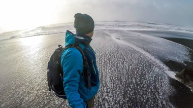 A young man wearing blue jacket and a beanie walks around black sand beach, with the sun behind him. Waves go high up on the beach. Rough and not calm sea.