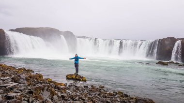 A young man standing at the a rock in front of an immense waterfall, surrounded by mountains, he spreads his arms wide. Edgy rock formation in the foreground. Stones overgrown with moss.