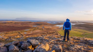 Man wearing a blue jacket, standing at the hill overlooking vast grassland and sea view. Wind blows his hoodie. Endless horizon, blending with sea line and mountains. Soft sunset colors.