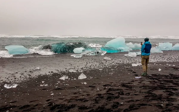 A young man wearing a blue jacket stands on black sand beach, holding his action camera and filming the rough sea washing the ice bergs to the shore. Blue shades of ice. Global warming effects.