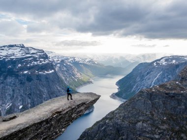 A man wearing blue jacket stands at the hanging rock formation, Trolltunga with a view on Ringedalsvatnet lake, Norway. Slopes of the mountains are partially covered with snow. Freedom and happiness