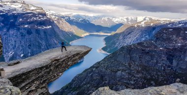A man wearing blue jacket stands at the hanging rock formation, Trolltunga with a view on Ringedalsvatnet lake, Norway. Slopes of the mountains are partially covered with snow. Freedom and happiness