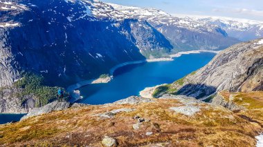 A man wearing blue jacket stands at the hanging rock formation with a view on Ringedalsvatnet lake, Norway. Slopes of the mountains are partially covered with snow. Bright day. Freedom and happiness