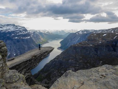 A man wearing blue jacket stands at the hanging rock formation, Trolltunga with a view on Ringedalsvatnet lake, Norway. Slopes of the mountains are partially covered with snow. Freedom and happiness