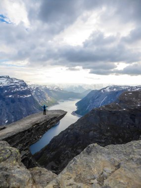 A man wearing blue jacket stands at the hanging rock formation, Trolltunga with a view on Ringedalsvatnet lake, Norway. Slopes of the mountains are partially covered with snow. Freedom and happiness
