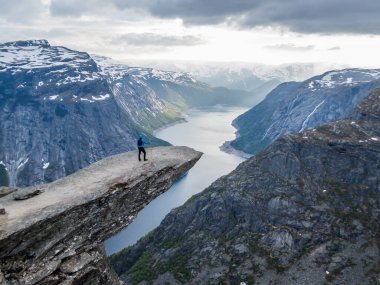 Mavi ceketli bir adam Norveç 'teki Ringedalsvatnet Gölü manzaralı Trolltunga' da asılı duruyor. Dağların yamaçları kısmen karla kaplı. Özgürlük ve mutluluk