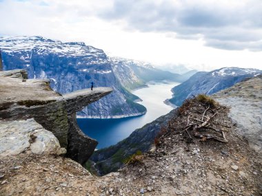 A man wearing blue jacket stands at the hanging rock formation, Trolltunga with a view on Ringedalsvatnet lake, Norway. Slopes of the mountains are partially covered with snow. Freedom and happiness