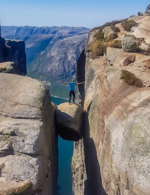 A young man wearing blue T-shirt standing on famous rock, Kjerag, Norway. The ball-shaped rock is hanging in between two stone walls with a free fall of 1000m. Boy spreads arms in a gesture of triumph