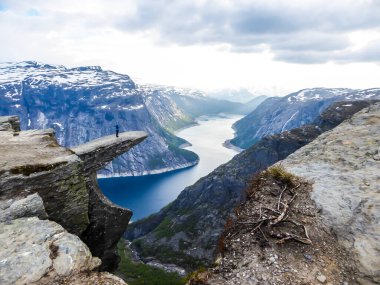 A man wearing blue jacket stands at the hanging rock formation, Trolltunga with a view on Ringedalsvatnet lake, Norway. Slopes of the mountains are partially covered with snow. Freedom and happiness