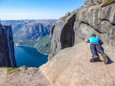 A young man lying at the edge of a steep mountain. He looks down into the valley. Overcoming the fear of heights. In front of her stunning Lysefjorden shimmering with many shades of blue and green