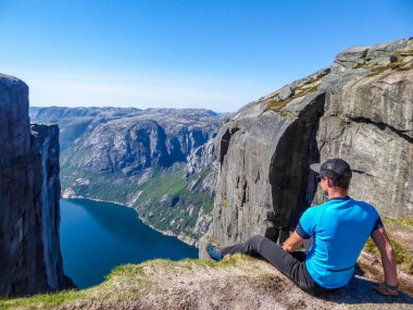 A young man sitting at the edge of a steep mountain. His legs are hanging down. Overcoming the fear of heights. In front of her stunning Lysefjorden shimmering with many shades of blue and green