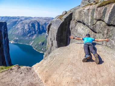 A man lying at the edge of a steep mountain pretends to be flying. He spreads arms wide in a gesture of freedom. In front of her stunning Lysefjorden shimmering with many shades of blue and green
