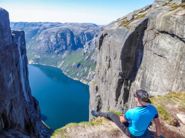 A young man sitting at the edge of a steep mountain. His legs are hanging down. Overcoming the fear of heights. In front of her stunning Lysefjorden shimmering with many shades of blue and green