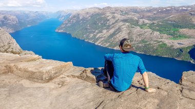 A man wearing blue shirt sits at the edge of a steep cliff of Preikestolen. A view on Lysefjorden. Fjord goes far inland. Man enjoys the view, feels free and happy. Great accomplishment