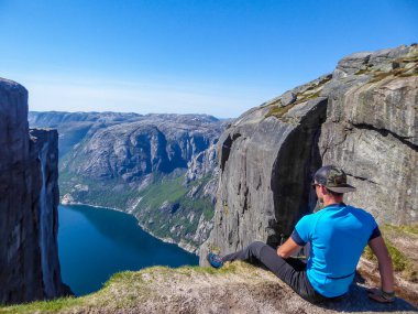 A young man sitting at the edge of a steep mountain. His legs are hanging down. Overcoming the fear of heights. In front of her stunning Lysefjorden shimmering with many shades of blue and green
