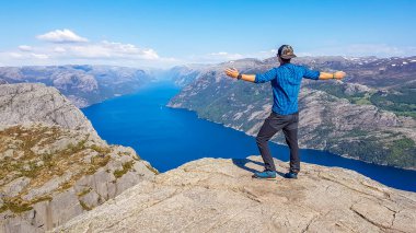 A man wearing blue shirt standsat the steep cliff of Preikestolen with his arms wide open. A view on Lysefjorden. Fjord goes far inland. Man enjoys the view, feels free and happy. Great accomplishment