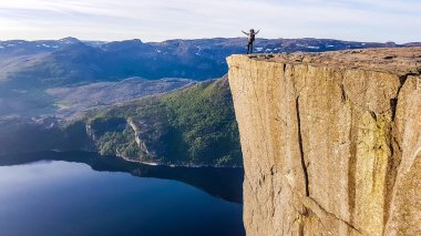 A boy with a huge hiking backpack standing at the edge of a steep cliff of Preikestolen, with a view on Lysefjorden. Fjord goes far inland. Boy enjoys the view, feeling free and happy.