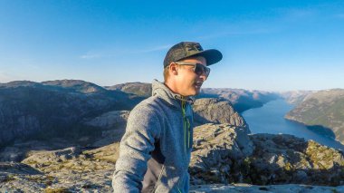 A young man in a hiking outfit walks along the edge of a steep cliff , with a selfie stick. He walks on a barren rocky cliffs. In the back is Lysefjorden, Norway. Bright and sunny day.