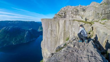A young man in a hiking outfit squats at the edge of a steep cliff with the view on Lysefjorden and famous pulpit rock - Preikestolen. Steep cliff going hundreds of meters down. Bright and clear day