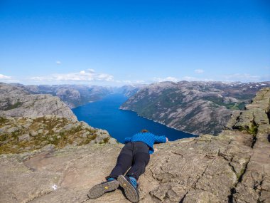 A man wearing blue T-shirt leaning over the edge of a steep cliff of Preikestolen, Pulpit Rock, with a view on Lysefjorden. Man is trying to overcome his fear of height. Overcoming acrophobia