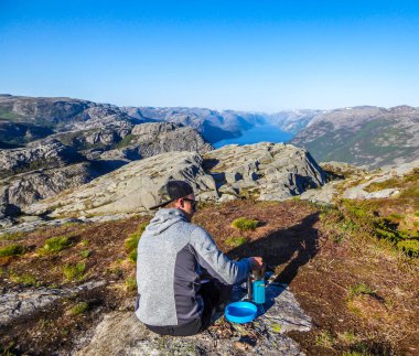 A young man preparing a dinner in the wilderness on a portable camping stove, with a view on the fjord. Blue plate next to the stove. A view on Lysefjorden. Fjord goes far inland. Soft sunset colors