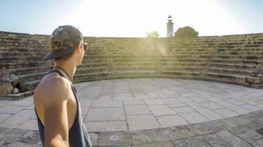 Young man taking a selfie in Kato Paphos Archaeological Park, ancient Greek and Roman city in Cyprus. Boy is wearing a full cap. Sun is shining bright. In the back a lighthouse is visible.
