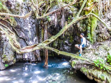 Young man wearing a full cap, admiring the Aphrodite's Bath, Cyprus. Small waterfall running down next to a steep rocks. Lush green flora. A tree branch hanging above the pond.
