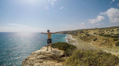 A young man standing at the steep cliff and spreading his arms wide in a gesture of freedom in Paphos, Cyprus. Blue sea spreads in front of him. Barren slopes of the cliff. Endless horizon line.