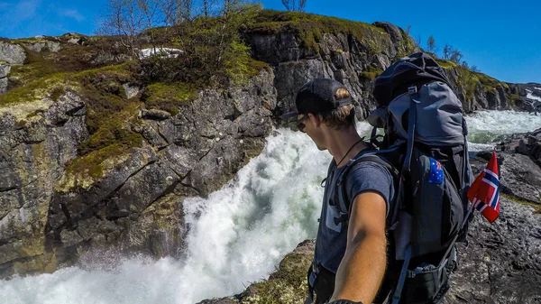A man stands next to a rough river in the highland village Vivelid, Norway and takes a selfie. The rough river flown is a gorge. The water is very foamy. Powerful current, slippery and dangerous rocks