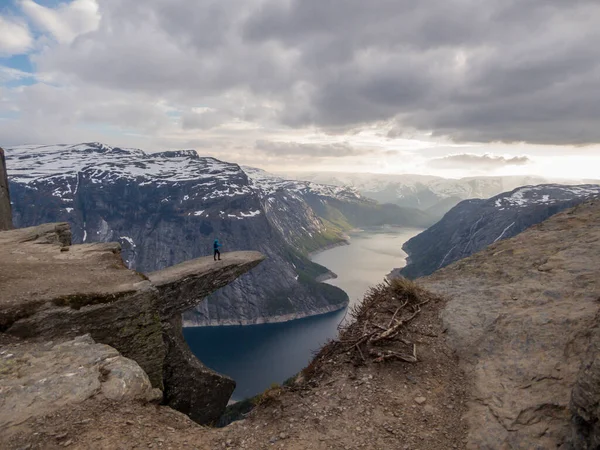 A man wearing blue jacket stands at the hanging rock formation, Trolltunga with a view on Ringedalsvatnet lake, Norway. Slopes of the mountains are partially covered with snow. Freedom and happiness
