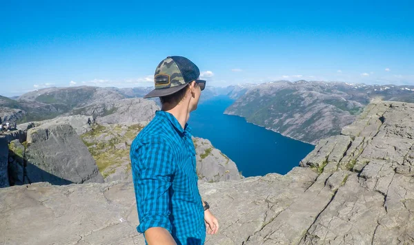A young man in a hiking outfit walks along the edge of a steep cliff , with a selfie stick. He walks on a barren rocky cliffs. In the back is Lysefjorden, Norway. Bright and sunny day.