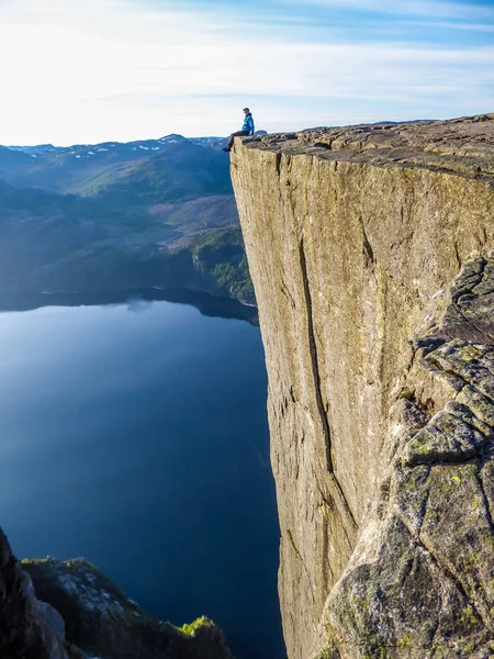 A man wearing blue shirt sits at the edge of a steep cliff of Preikestolen with his legs hanging down. A view on Lysefjorden. Fjord goes far inland. Man enjoys the view, feels free and happy.