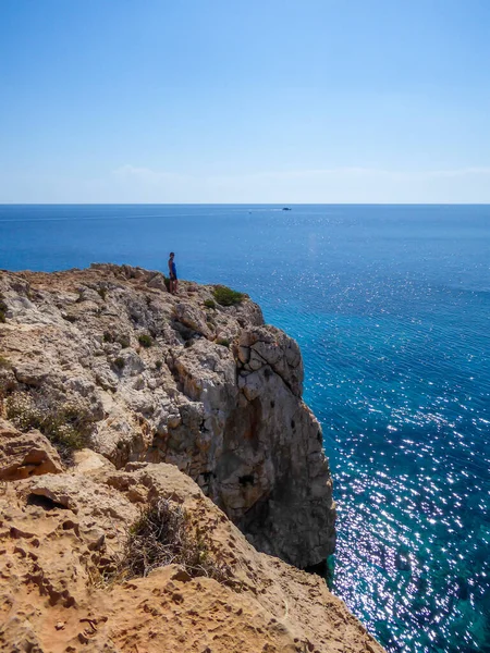 A young man standing at the steep cliff of Cape Greco, Cyprus. The cliff looks dangerous. Barren slopes of the stony formation. Sea has many shades of blue. Endless line of a horizon. Clear day.