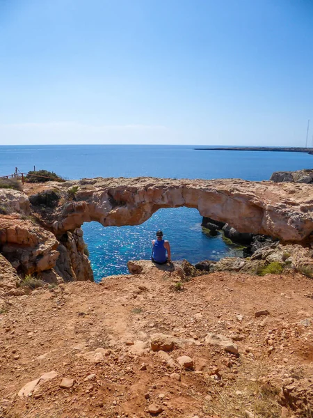 A young man sitting at the steep cliff of Cape Greco, Cyprus, in front of his is a stony arch, bridge connecting two cliffs. Barren slopes of the stony formation. Blue sea. Endless line of a horizon.