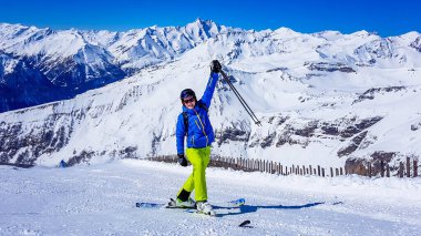 A skier standing on the side of a slope in Heiligenblut, Austria. Perfectly groomed slopes. High mountains surrounding the man wearing yellow trousers, blue jacket and helm. He throws his hands up.