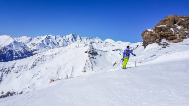 A skier standing on the side of the piste, enjoying the view. Perfectly groomed slopes. High mountains surrounding the man wearing yellow trousers and blue jacket. Man wears helm for the protection.