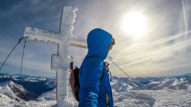 A man in skiing outfit standing on the top of a mountain peak with a snow covered cross behind him. Endless snow caped mountains around the man. Soft clouds on the sky. Moelltaler Glacier in Austria