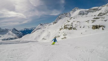 A skier going down the slope in Moelltaler Gletscher, Austria. Perfectly groomed slopes. High mountains surrounding the man wearing yellow trousers and blue jacket. Man wears helm for the protection.