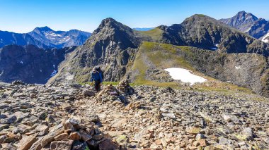 A young man with a big backpack hiking up a steep and stony trail in high Alps, Schladming region in Austria. There are endless mountain ranges around him. Some snow on the slopes. Clear sky.