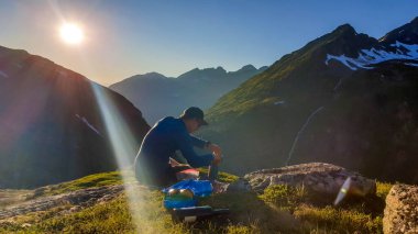 A young man cooking a dinner in the wilderness during the sunrise. He is sitting at the edge of a mountain, mixing the dish. He is surrounded by tall mountains. Alpine camping. Outdoor kitchen.