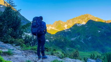 A man with a big backpack standing on the trail and looking at the first sunbeams touching the tall mountain peaks. Calm and serene morning in Alps. Schladming region, Austria.
