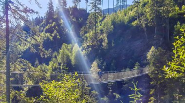A young man standing on a suspension bridge that hangs high above the gorge. Both slopes of the mountains are overgrown with thick forest. Sunbeams shining bright. Clear day.
