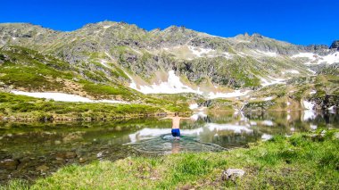 A young man walking into the icy-cold alpine lake. Bravery and resistance to cold temperatures. There is a lot of snow around on the mountain's slopes.Spring is slowly coming into the valley.